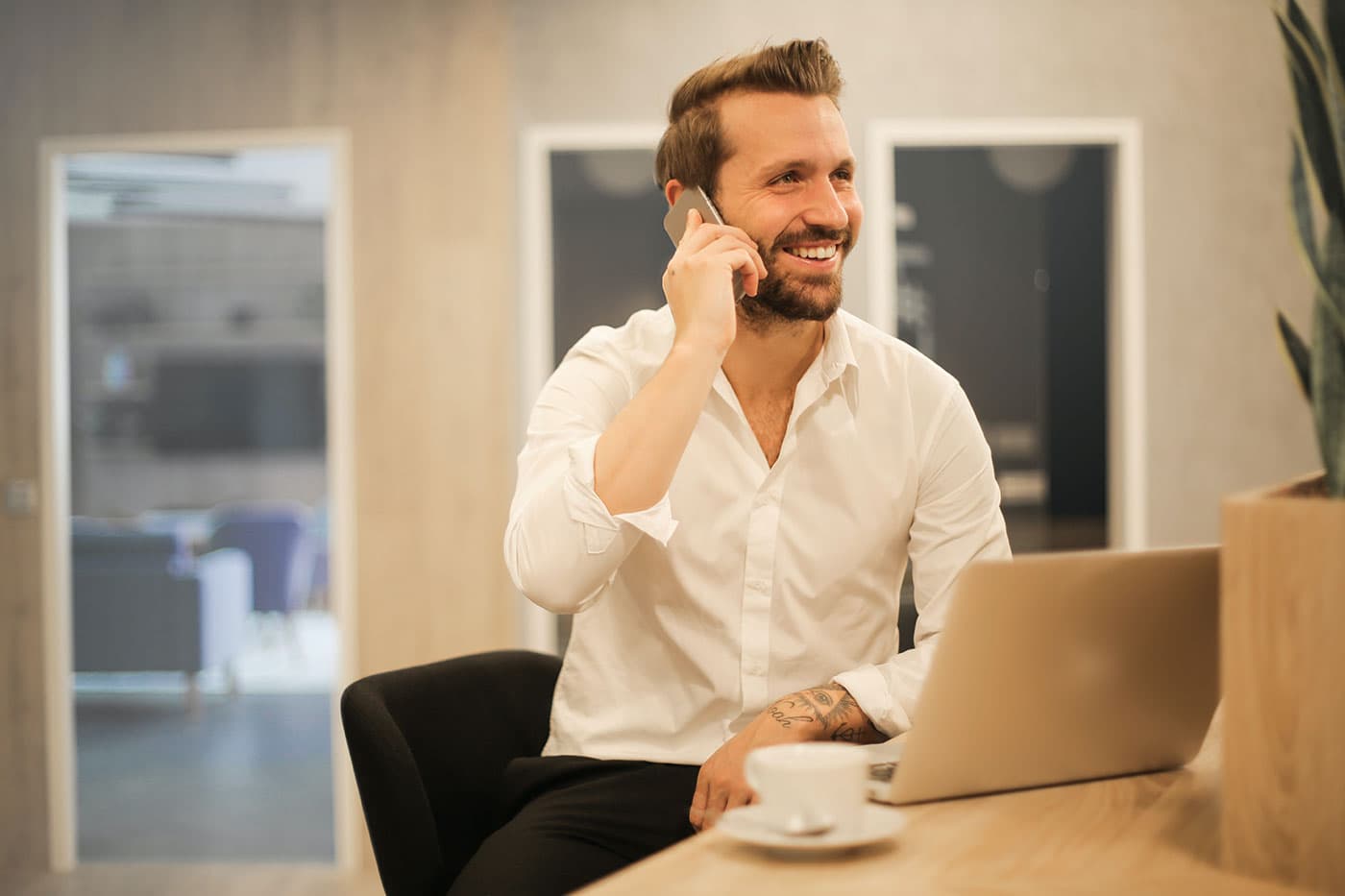 Soutien Professionnel et Communication Portrait d'un homme d'affaires souriant et barbu, parlant au téléphone portable à un bureau avec un ordinateur portable et une tasse de café, illustrant la communication professionnelle.