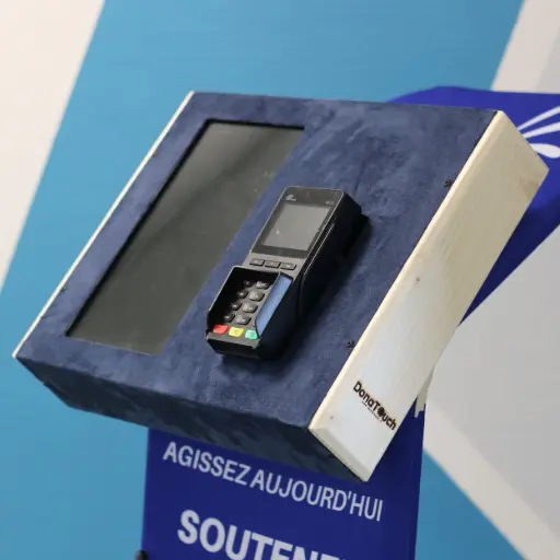 Gros plan sur un pupitre de don DonaTouch (ou iTouchAm) avec un revêtement en suédine bleue et un panneau latéral en bois clair, montrant un terminal de paiement électronique prêt à l'emploi.