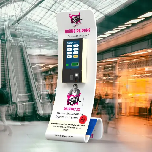 Borne de dons DonaTouch aux couleurs des Restos du Cœur (blanc et rouge), affichant le message "BORNE DE DONS - On compte sur vous !" dans un hall de gare ou d'aéroport.